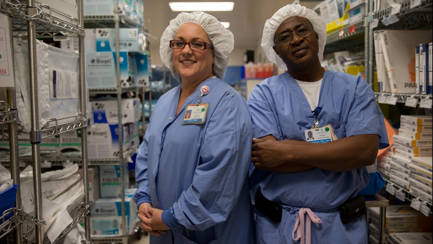Two health care workers, wearing blue scrubs and white hair coverings, in a supply closet