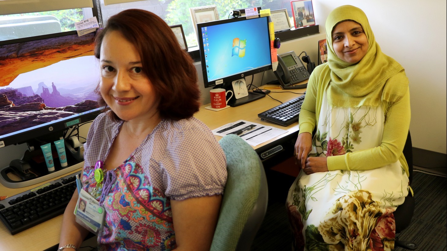 Two women, one wearing a yellow hijab, at a desk with a computer, smiling