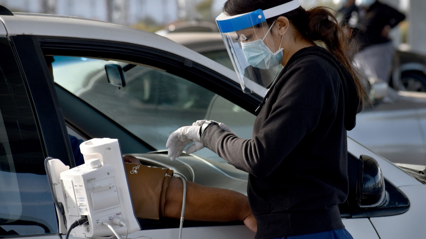 Woman wearing face shield checking a man's blood pressure while he sits in the passenger seat of a car