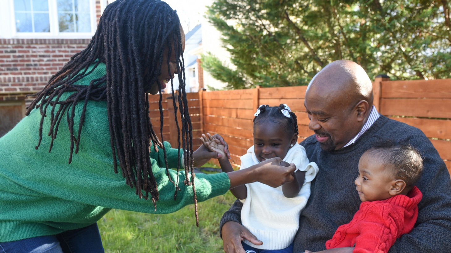 In a sunny backyard, a Black mom holds hands with her young daughter, who is sitting with her brother in the lap of their dad.