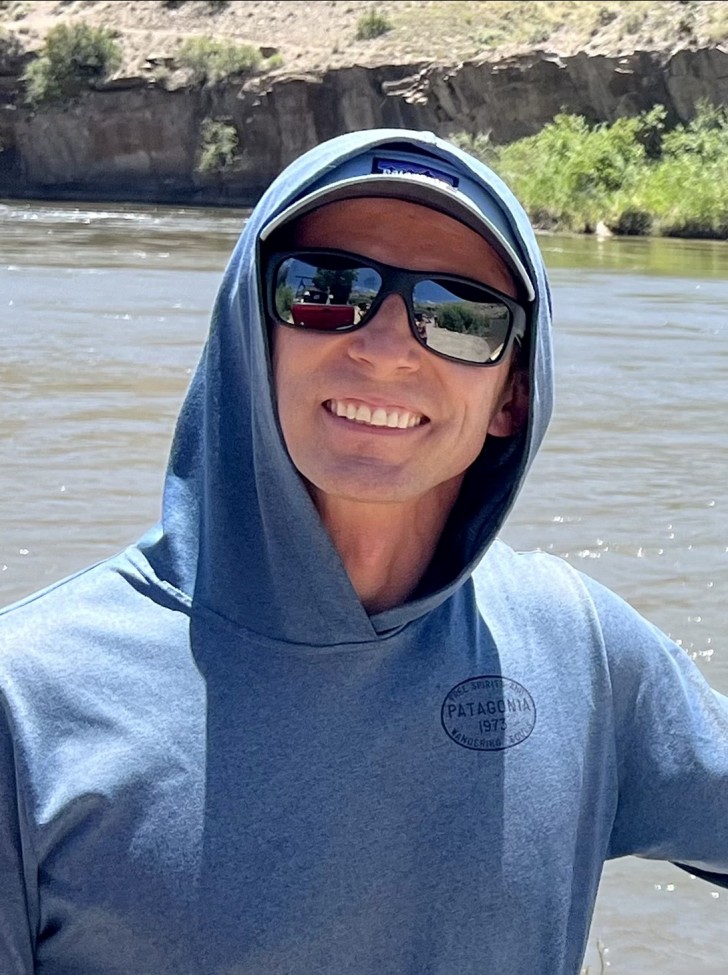 a young white man, wearing a blue hoodie and sunglasses, in front of a river and smiling